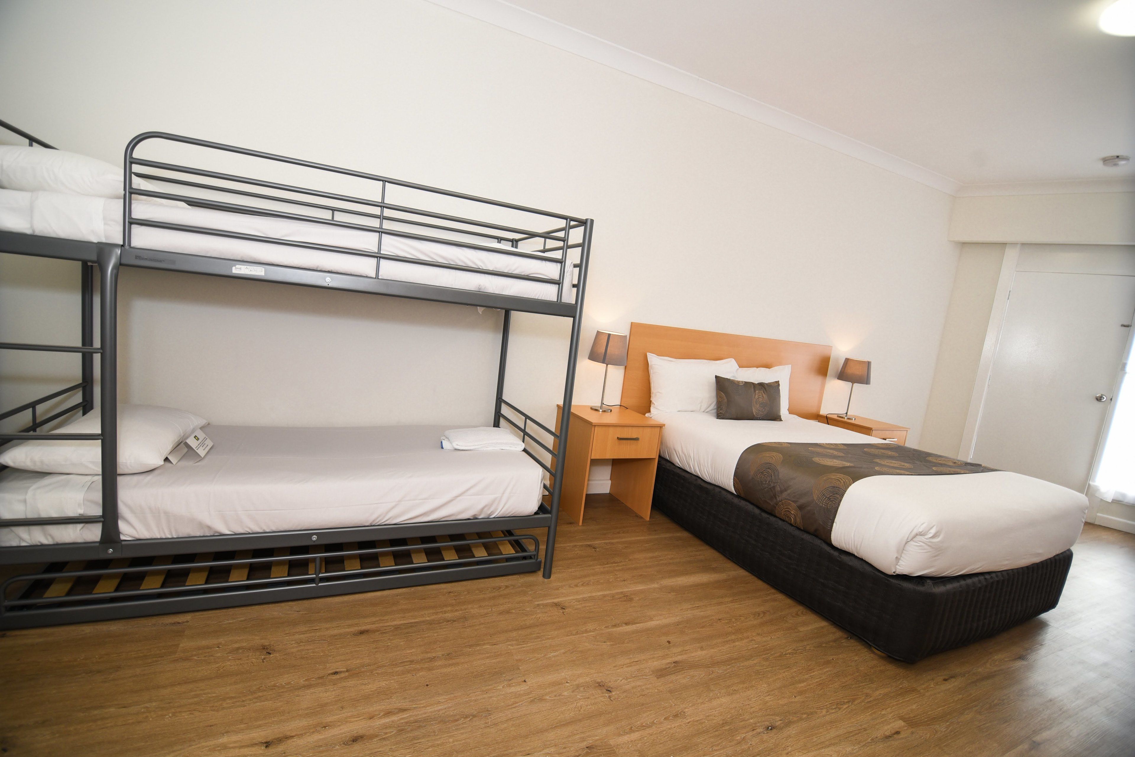 A room with bunk beds, a single bed, wooden flooring, a nightstand with a lamp, and a door.
