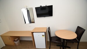 Una stanza compatta con una scrivania in legno, un piccolo frigorifero bianco, un tavolino rotondo con due sedie, una TV a parete e uno specchio.