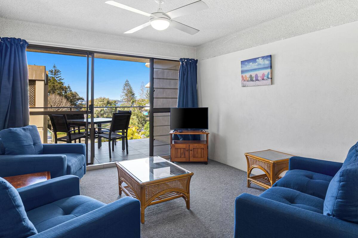 A room with a balcony, blue sofas, a glass coffee table, a television on a wooden cabinet, and a painting on the wall.