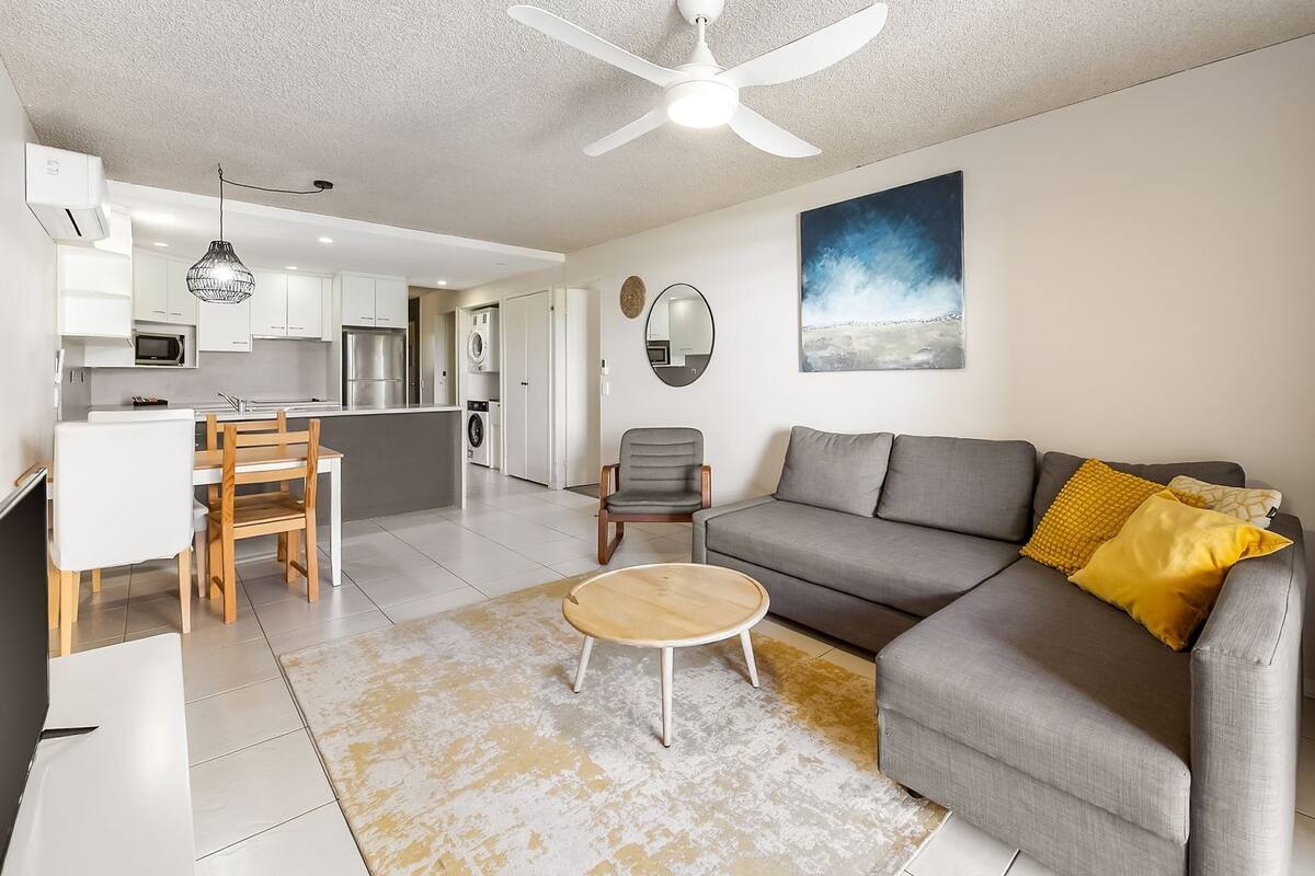 A modern living room with a grey sofa, a wooden coffee table, and a dining area with white chairs and a white table.