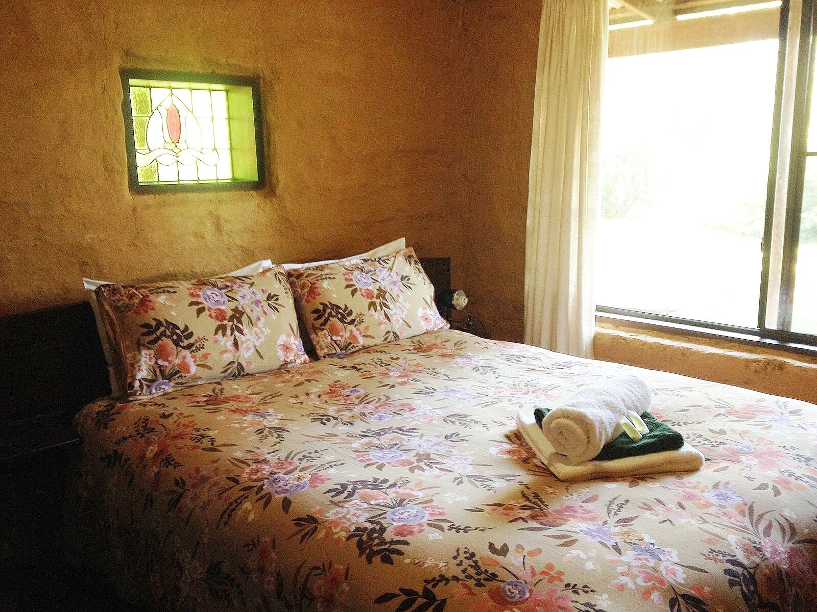 A bedroom with a floral bedspread, two matching pillows, a small window with a green frame, and a window with curtains.