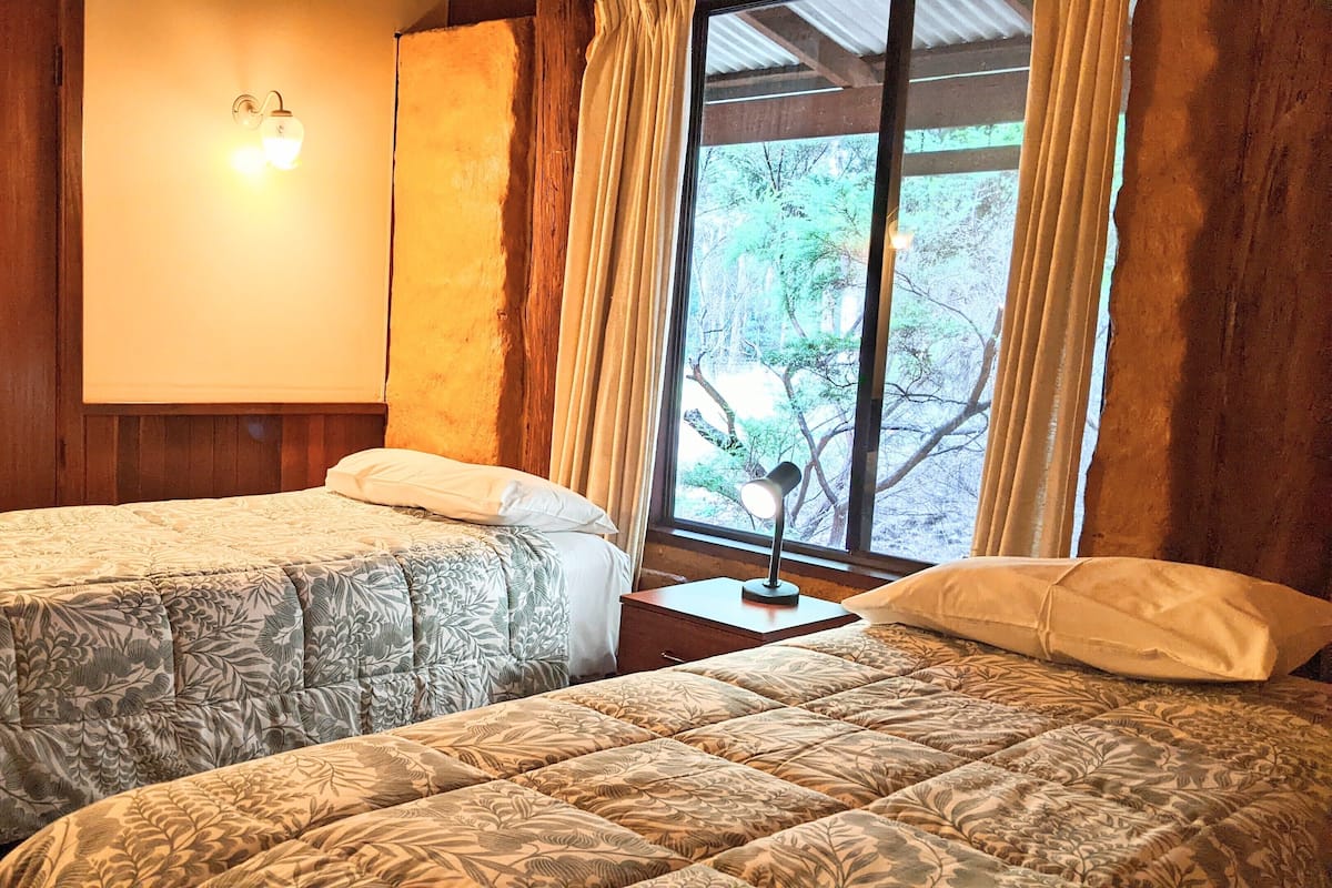 A room with two beds, wooden walls, a window with curtains, and a wall-mounted light fixture.