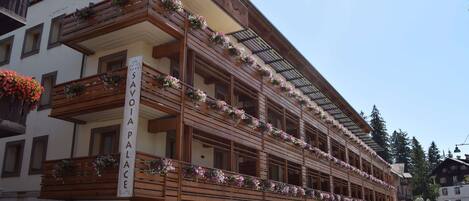 Un hotel de varios pisos con balcones decorados con flores, un cartel que dice "SAVIA PALACE" y un estilo arquitectónico clásico.