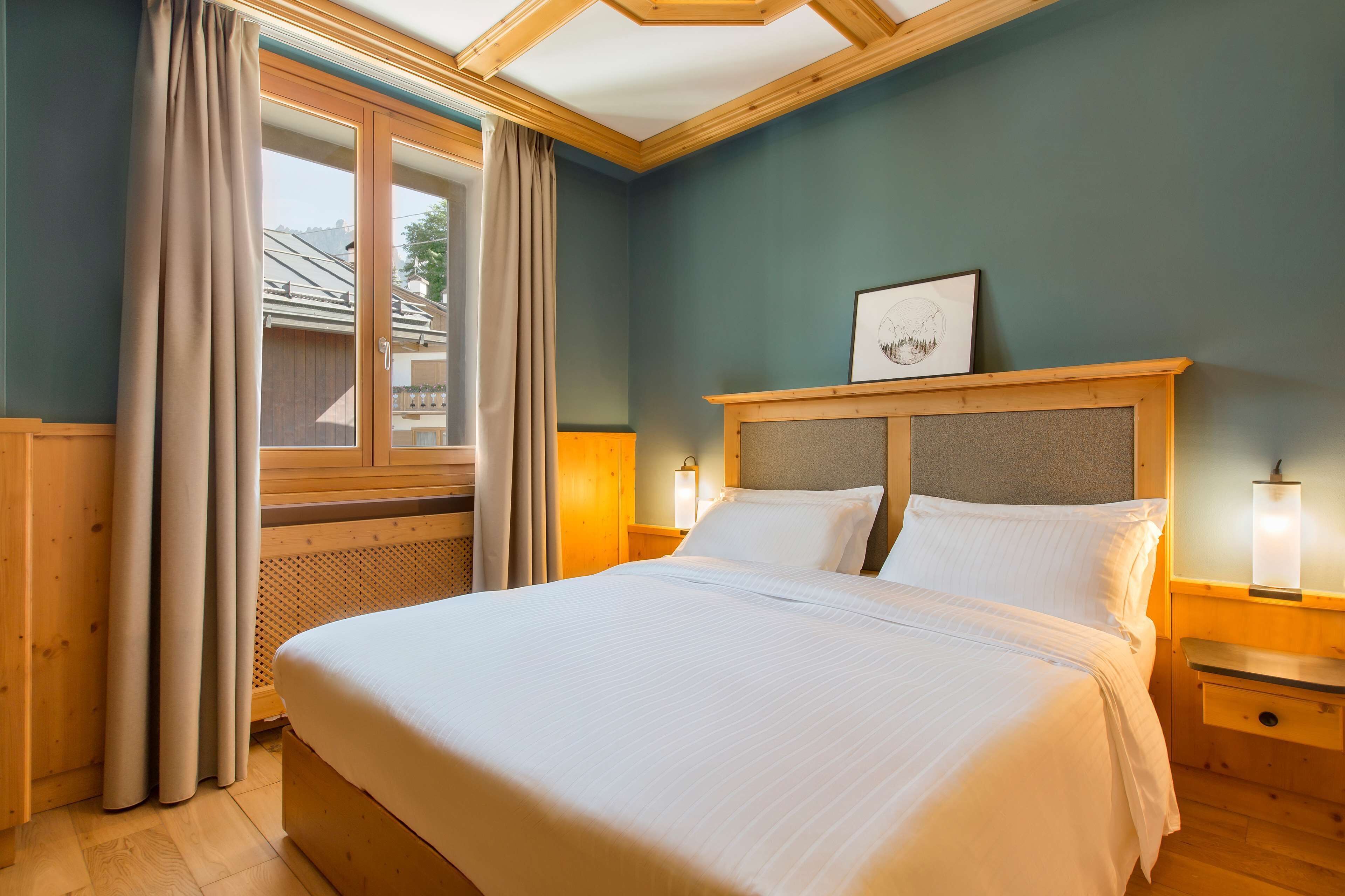 Ein ordentlich bezogenes Bett in einem Zimmer mit einem hölzernen Kopfteil, einem Fenster mit Vorhängen und einem gerahmten Bild an der Wand.