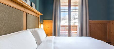 A hotel room with a large bed, a wooden headboard, a window with curtains, and a view of a snowy landscape.
