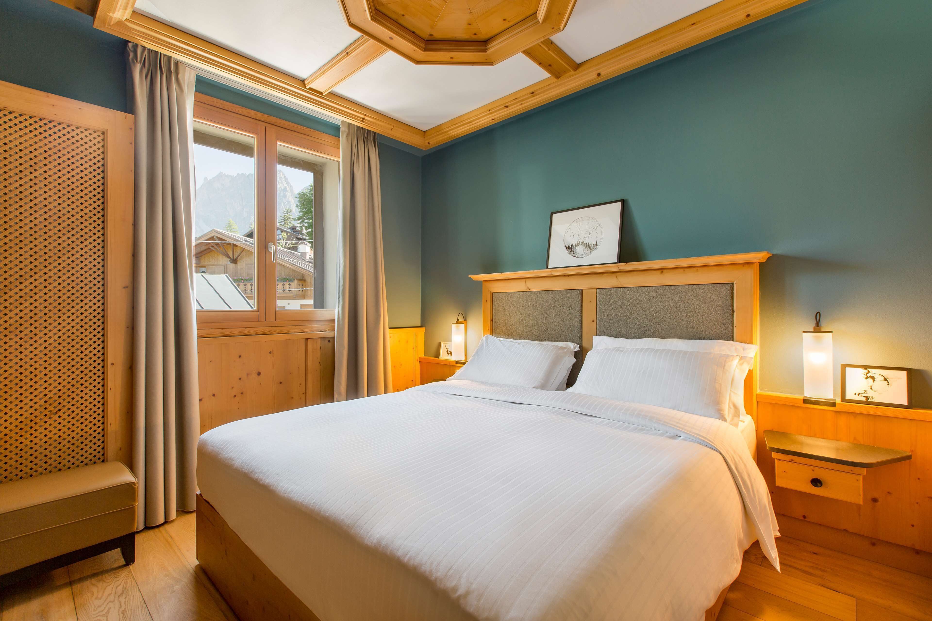 Un dormitorio con una cama grande, cabecera de madera, dos mesitas de noche con lámparas, una ventana con cortinas y vistas a montañas.