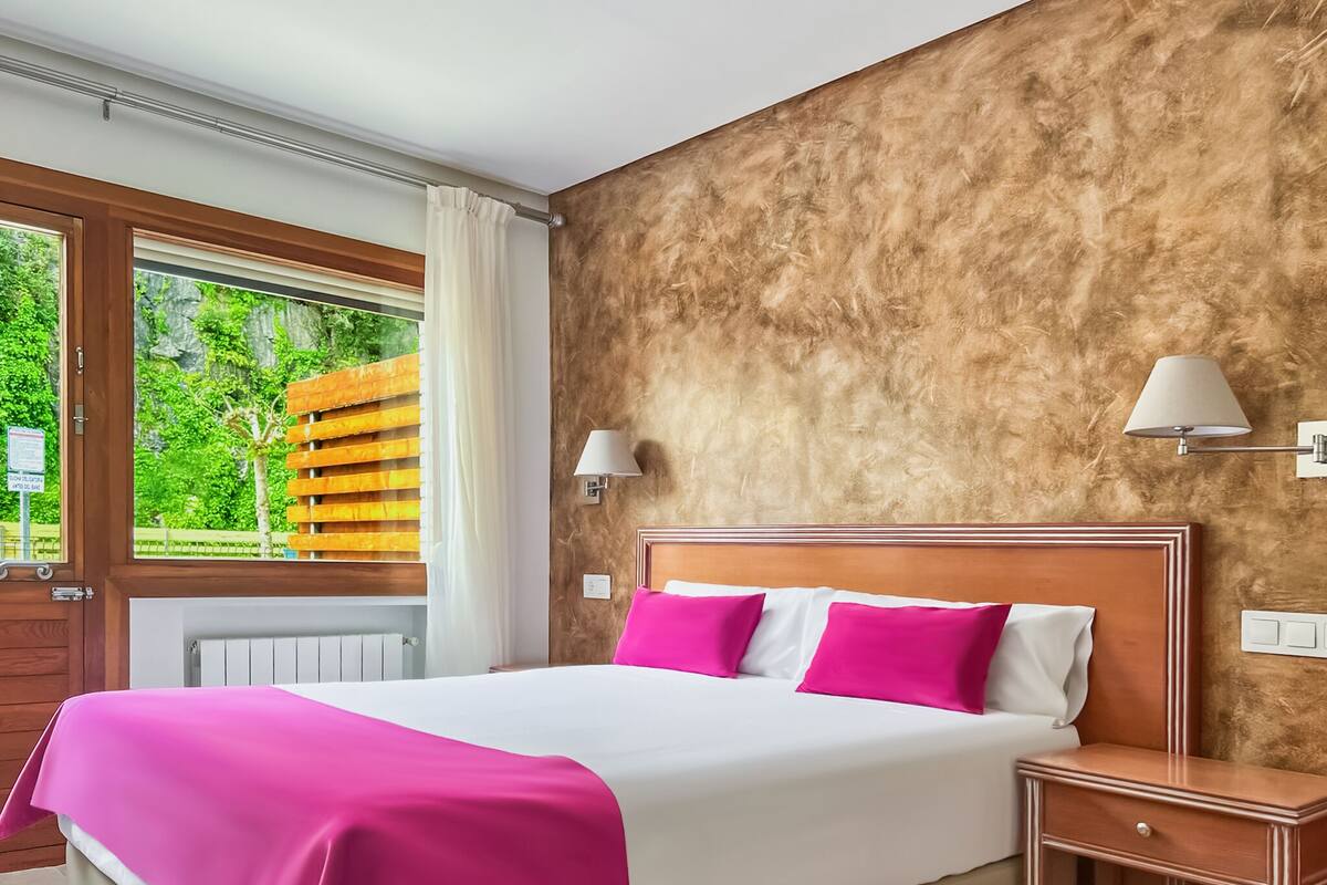 Ein Hotelzimmer mit einem großen Bett, einem Holzkopfende, zwei Nachttischlampen, einem Nachttisch und einem Fenster mit Blick auf Grünflächen.