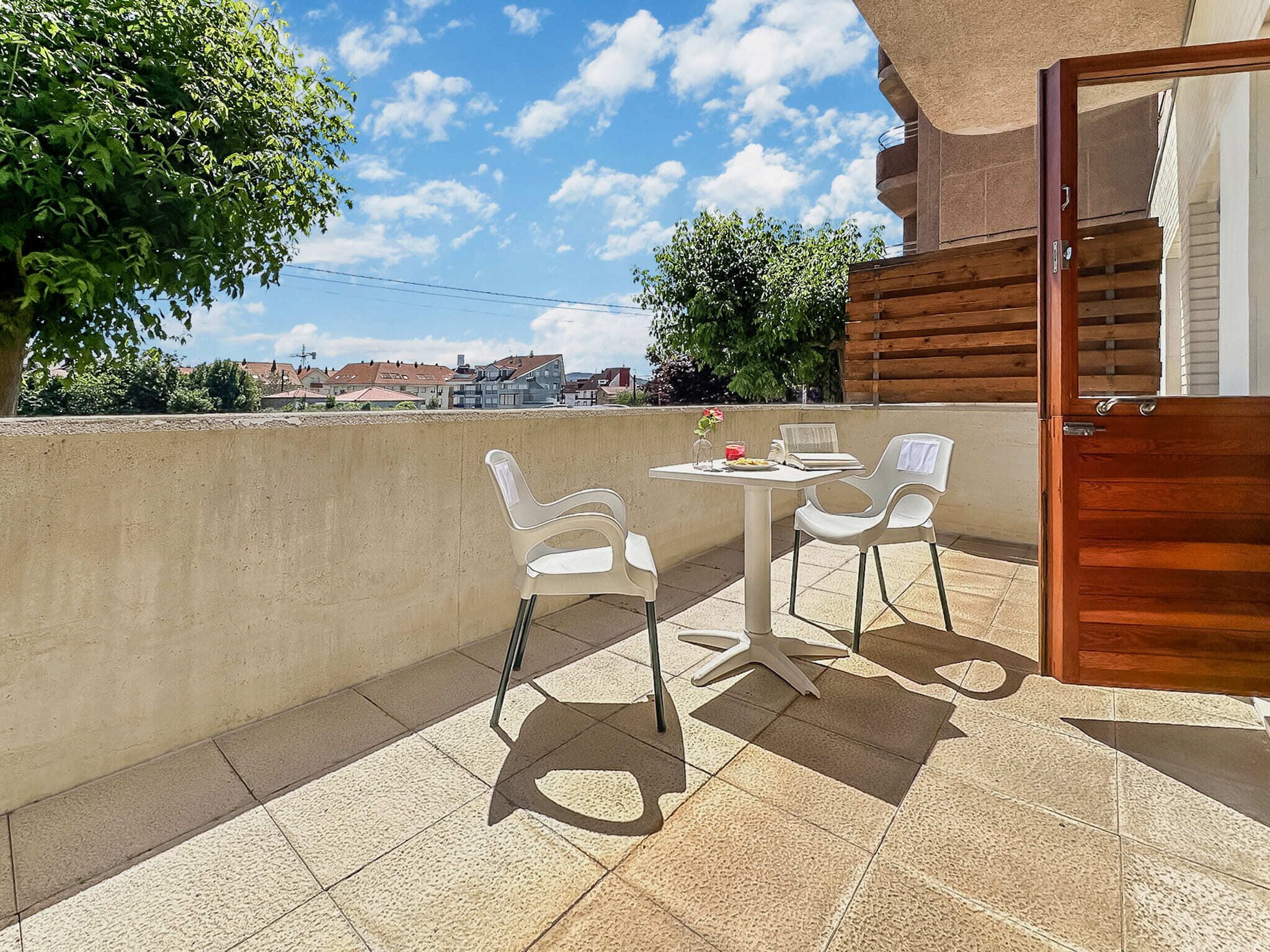 Un balcón con mesa y sillas, una puerta de madera y vistas a edificios al fondo.
