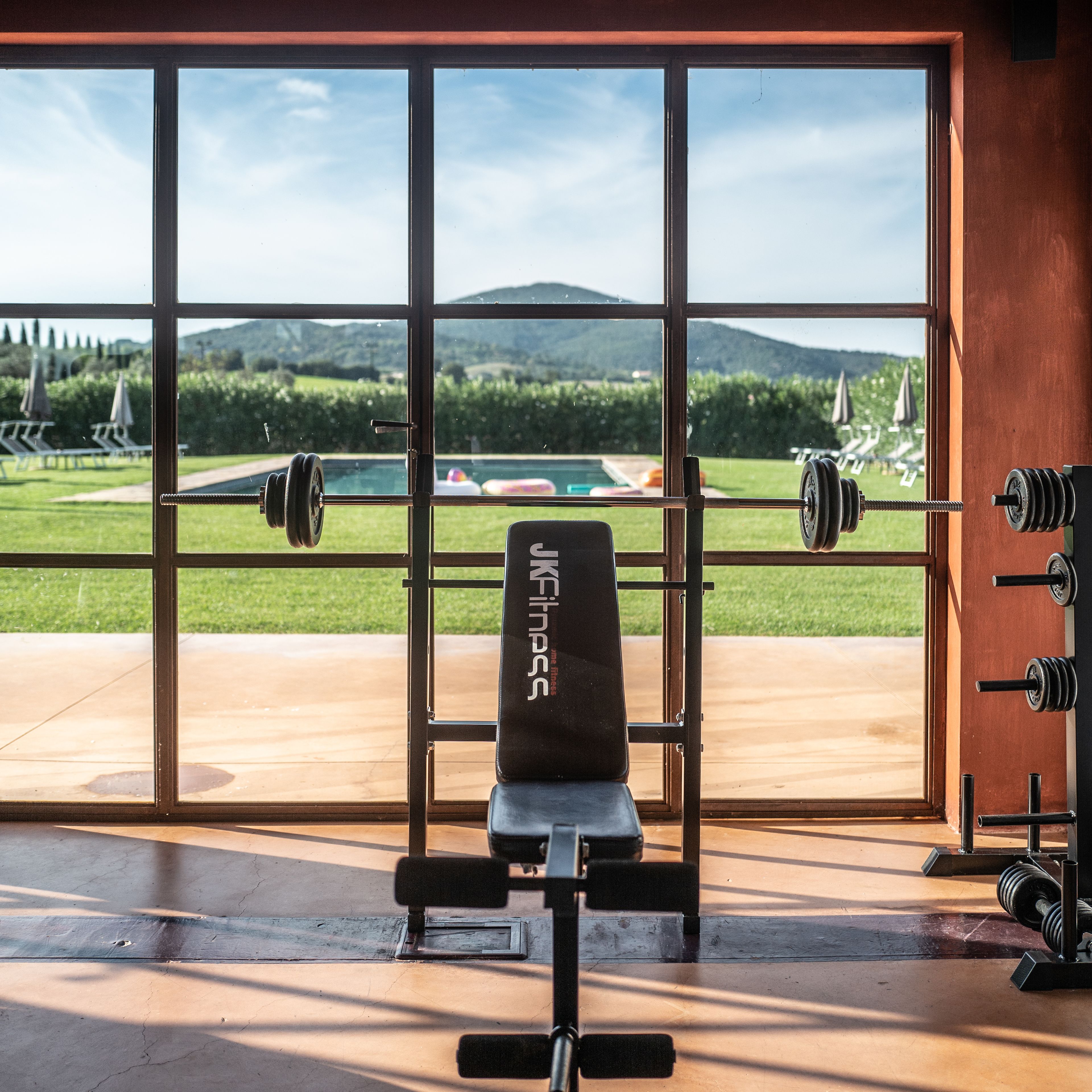 A gym with a bench and weight racks, large windows with a view of a pool and mountains.