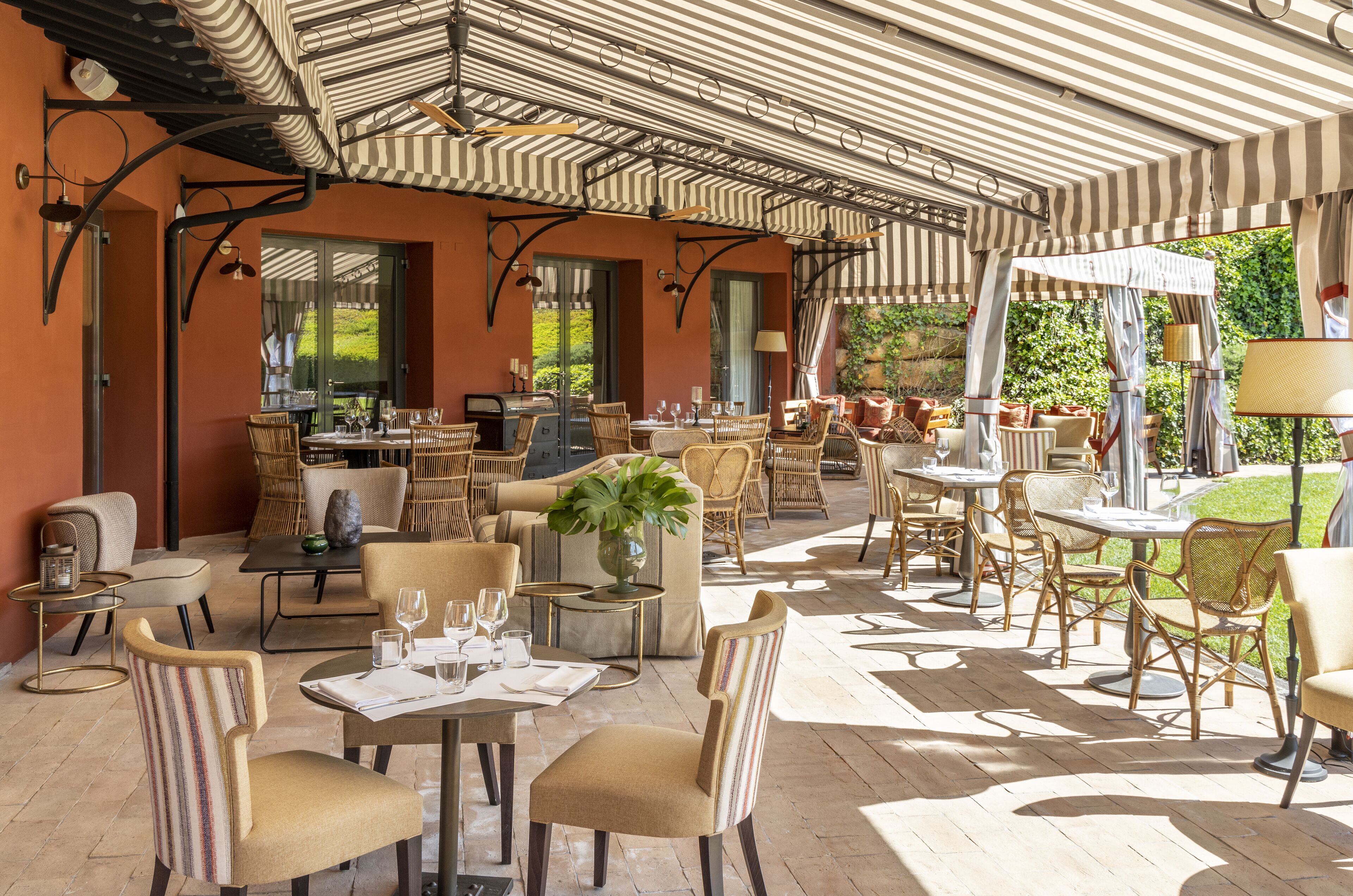 Eine Terrassenfläche im Freien mit Korbmöbeln, Tischen und einem Streifenverdeck.