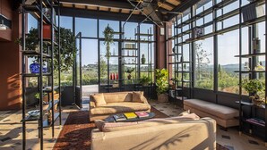 A modern living room with a beige sofa, a coffee table, and a bookshelf. Large windows provide a view of the outdoors. - Resort Locanda Rossa (Capalbio)