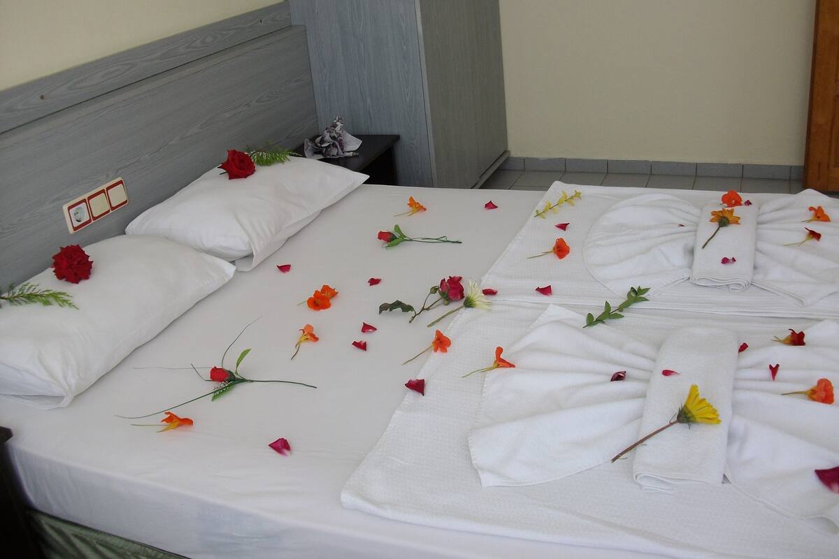 A neatly made bed with white linens, a red rose, and scattered flower petals.