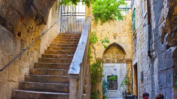 A narrow stone staircase leading to an arched doorway labeled "ISPOD URE" with outdoor seating and potted plants. - Rodrigo Apartments (Split)