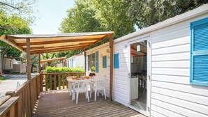 Une terrasse en bois avec une table et des chaises, un espace couvert, et une maison mobile blanche avec des volets bleus.