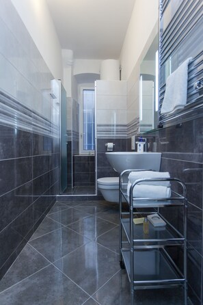A modern bathroom with dark tiles, a glass shower enclosure, a white sink, and a metal towel rack.