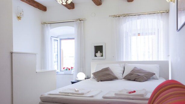 A neatly made bed with white linens and pillows, a bedside table with a lamp, and a window with curtains. - Dubrovnik Center Apartments (Dubrovnik)