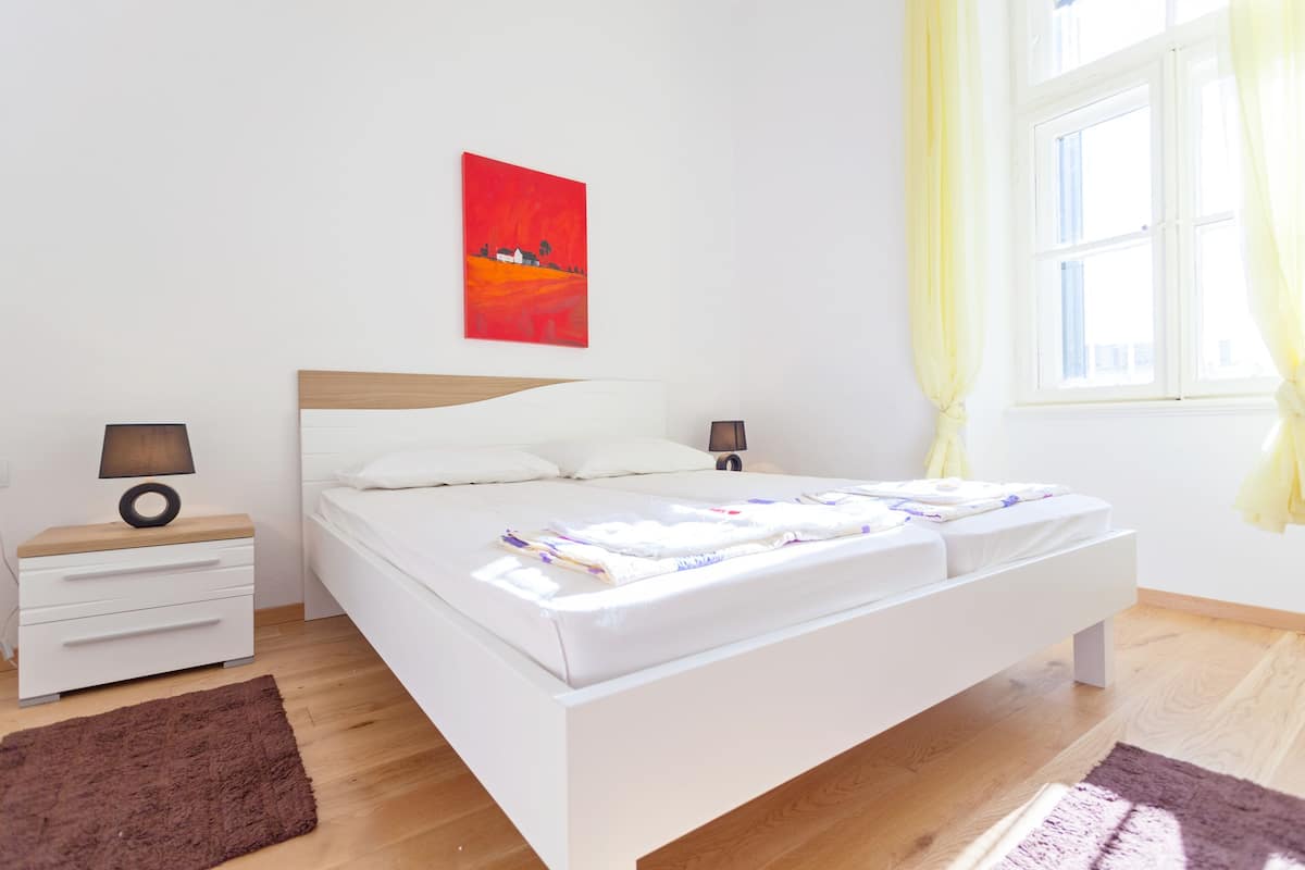 A modern bedroom with a large bed, bedside tables, a nightstand, a window with curtains, and a painting on the wall.
