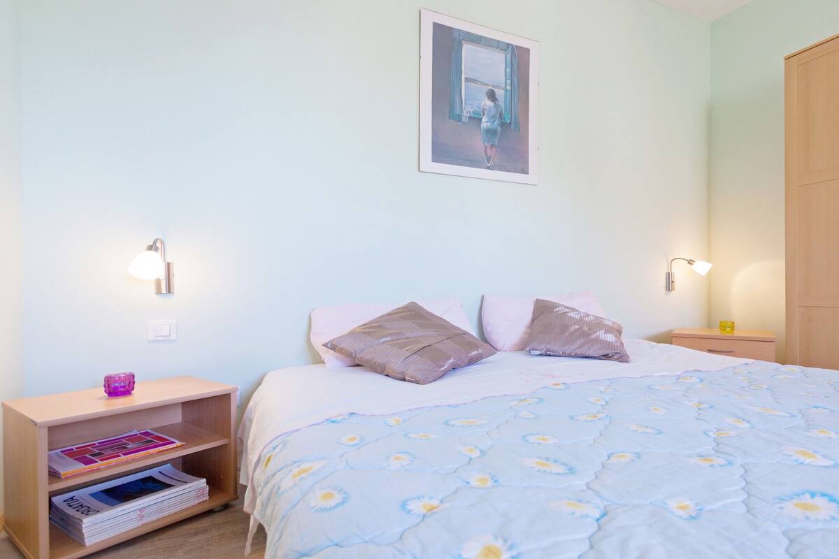 A bedroom with a bed, two bedside tables, a nightstand, a wardrobe, and a wall-mounted picture.