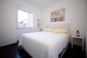 A bedroom with a large bed, a window, a bedside table with a lamp, and a colorful abstract painting on the wall. - Dubrovnik Center Apartments (Dubrovnik)