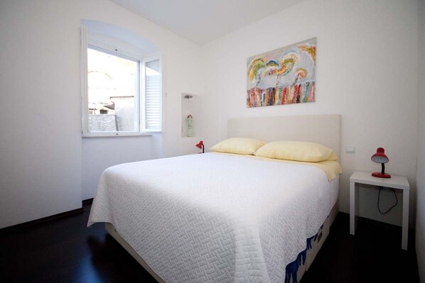 A bedroom with a large bed, a window, a bedside table with a lamp, and a colorful abstract painting on the wall.