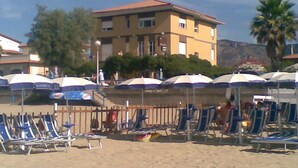 A beach with sun loungers and umbrellas, a building in the background, and a palm tree. - Hotel San Carlo (Terracina)