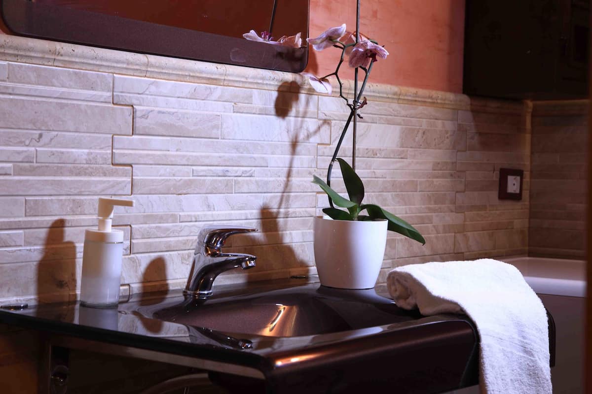 A bathroom with a marble wall, a sink with a faucet, a potted orchid, and a towel on the edge of a bathtub.