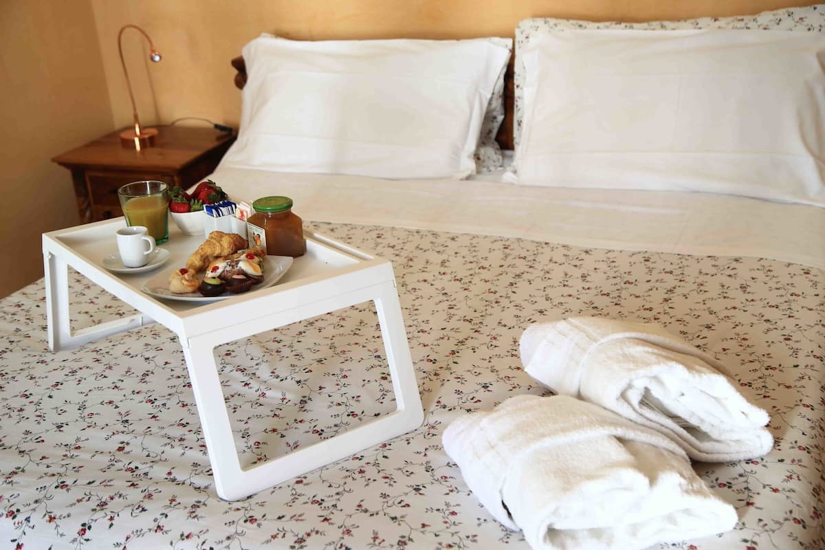 A neatly made bed with white linens and pillows, a small bedside table with a tray of breakfast items including toast, a cup, and a glass of juice, and a pair of folded white towels on the bed.