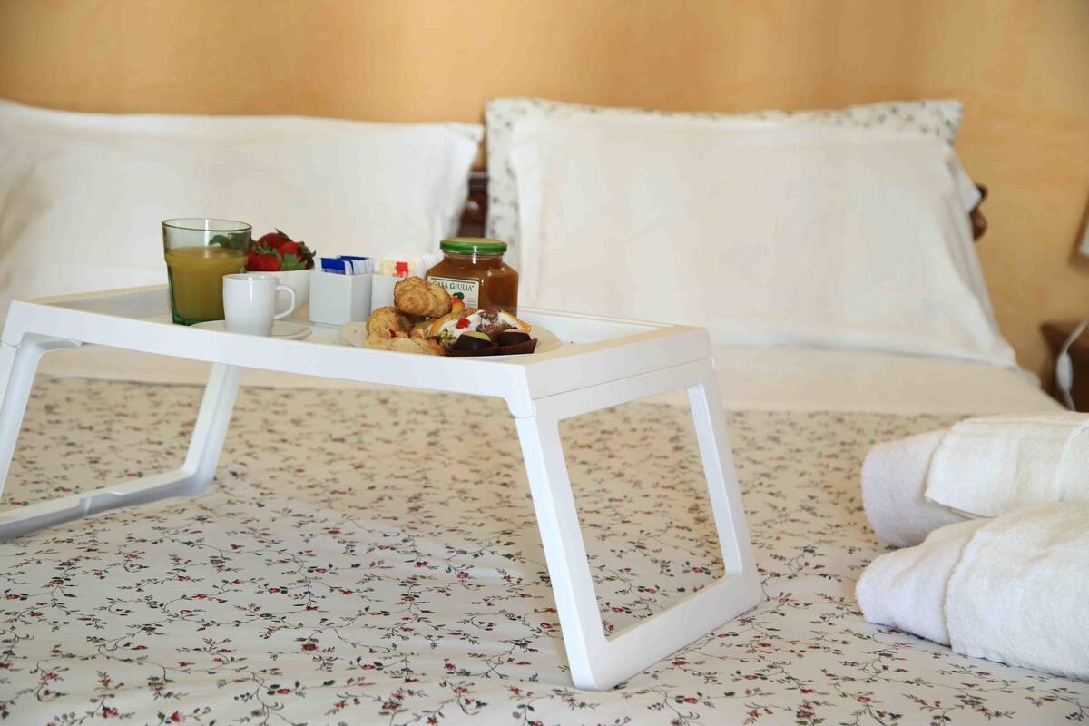 A neatly made bed with a floral bedspread, a white bedside table with breakfast items, and a white pillow.