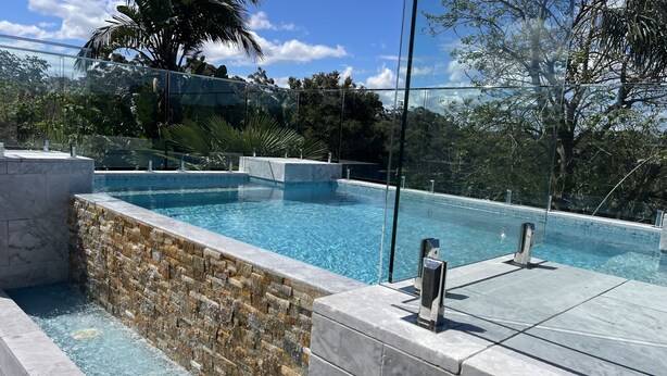 A modern infinity pool with clear blue water, surrounded by stone walls and glass railings, set against a backdrop of lush greenery and a clear sky.