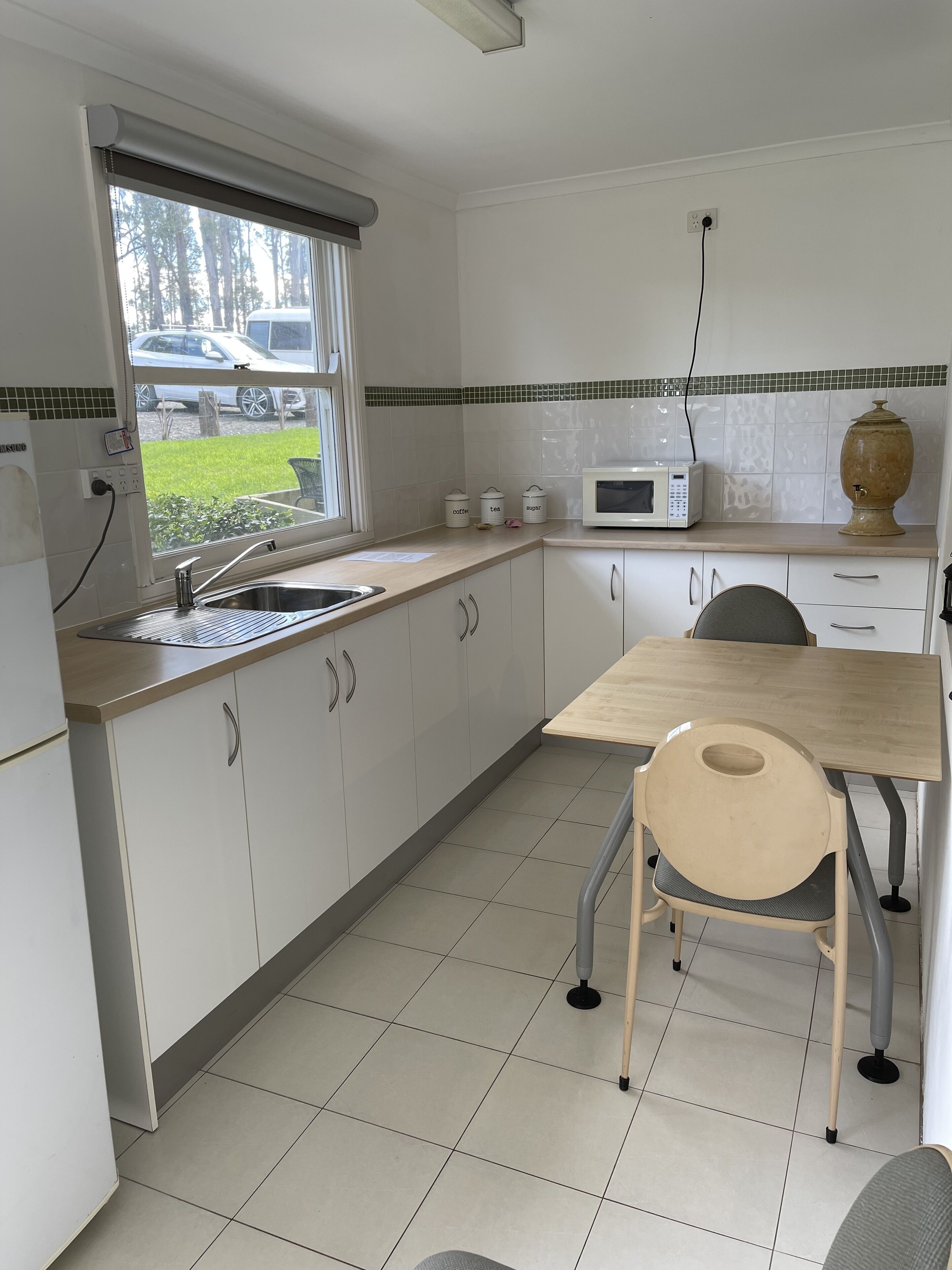 A kitchen with white cabinets, a microwave, a sink, and a window with a view of parked cars and trees.