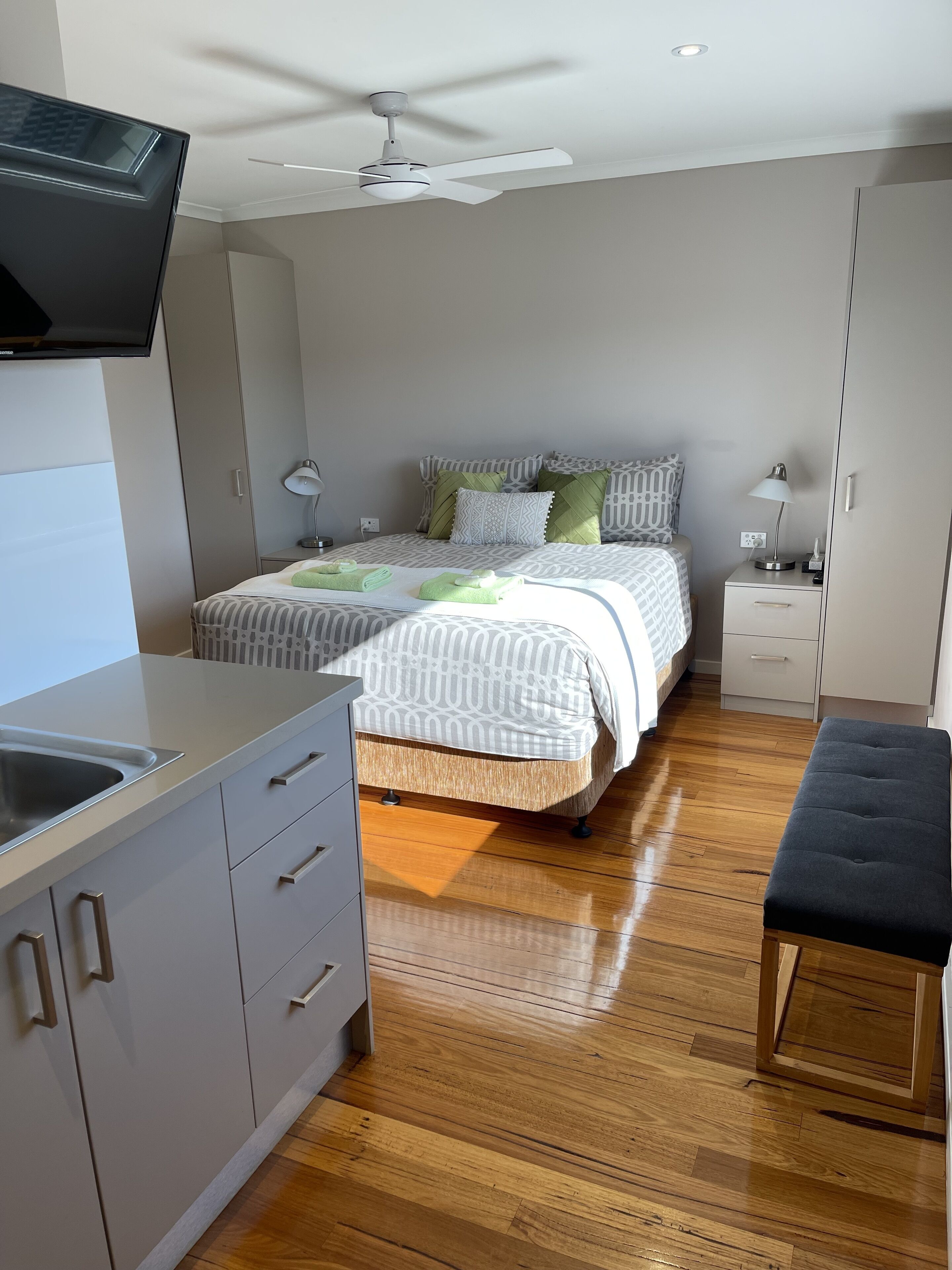 A modern bedroom with a bed, bedside tables, a bench, a ceiling fan, and a TV.