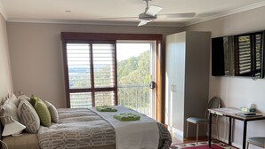 A bedroom with a bed, a desk, a chair, a TV, and a balcony with a view.