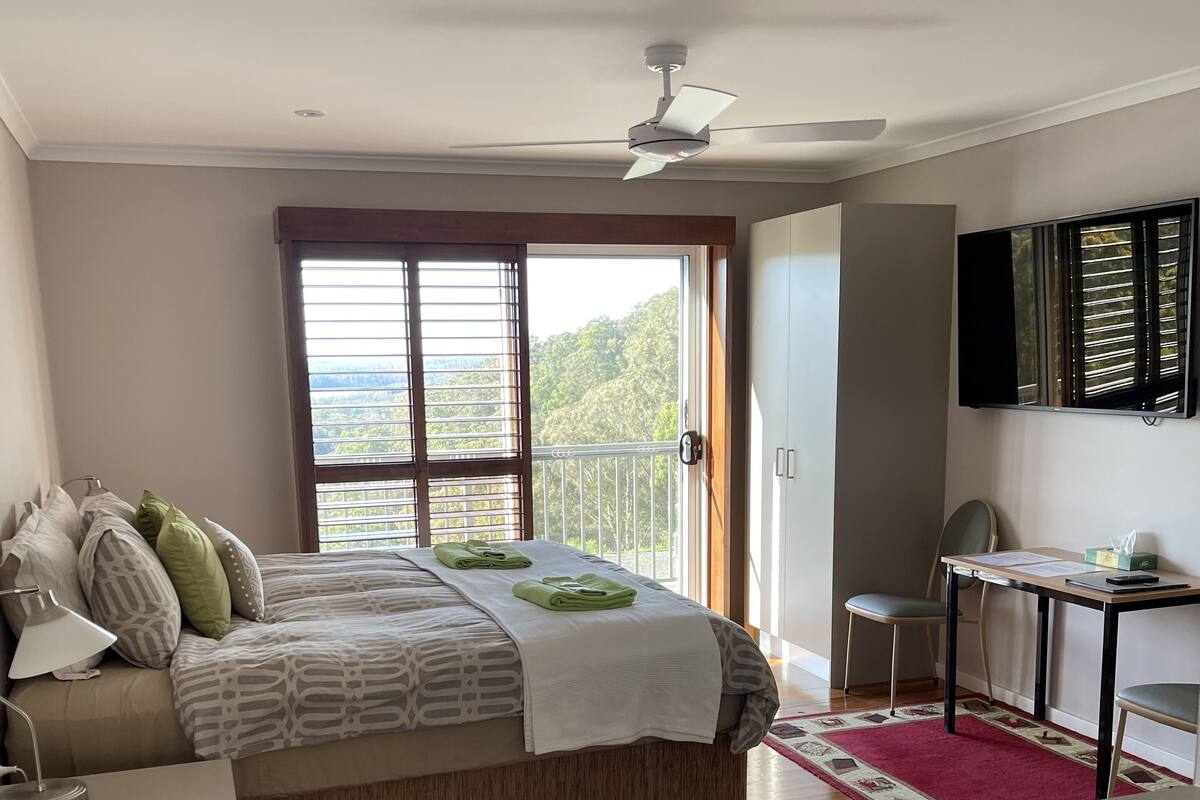 A bedroom with a bed, a desk, a chair, a TV, and a balcony with a view.