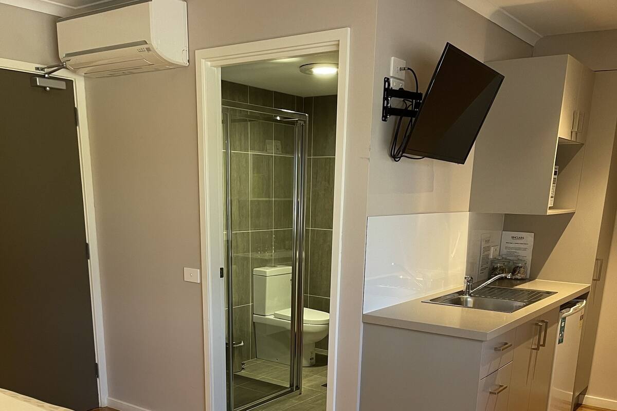 A compact hotel room with a bed, a small kitchenette, a shower, and a toilet.