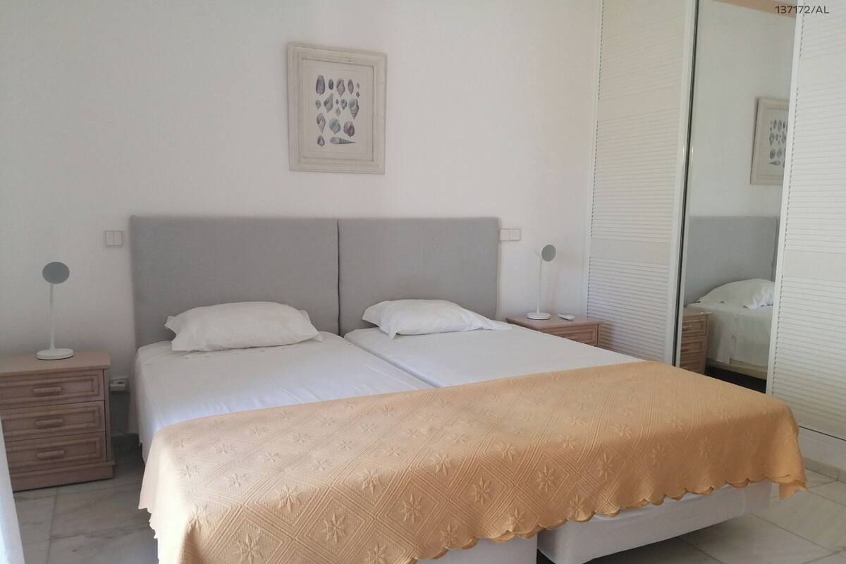 A double bed with a beige blanket, two bedside tables with lamps, a framed artwork on the wall, and a sliding door closet.