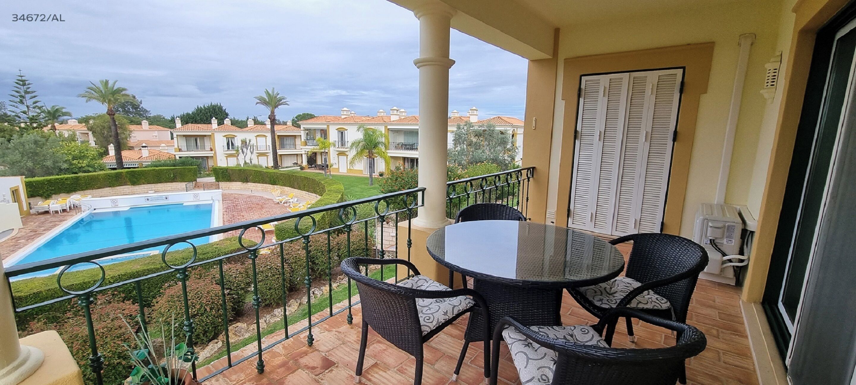 A balcony with a glass table and chairs, overlooking a swimming pool and a well-maintained garden.