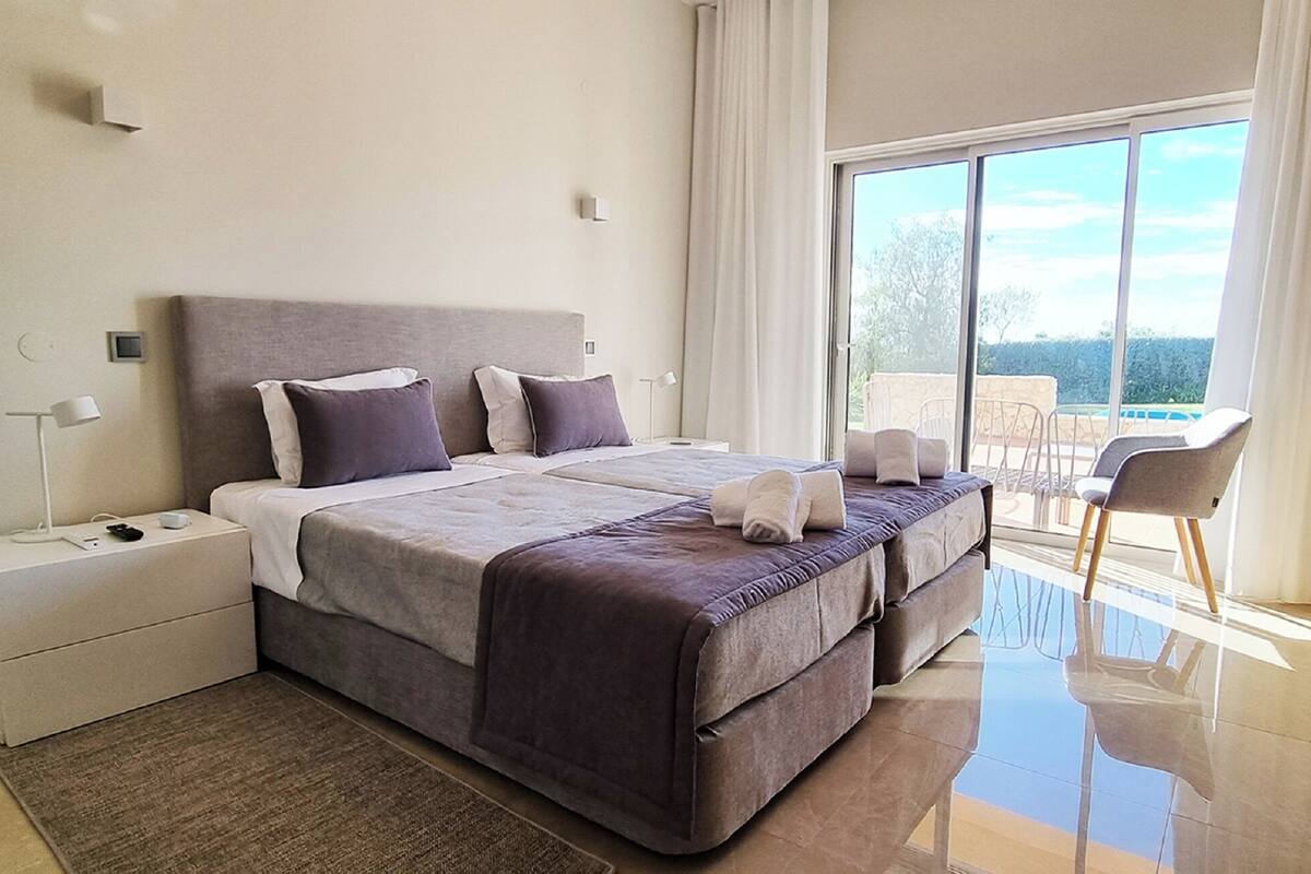 A modern bedroom with a large bed, a nightstand, a chair, and a view of the outdoors.