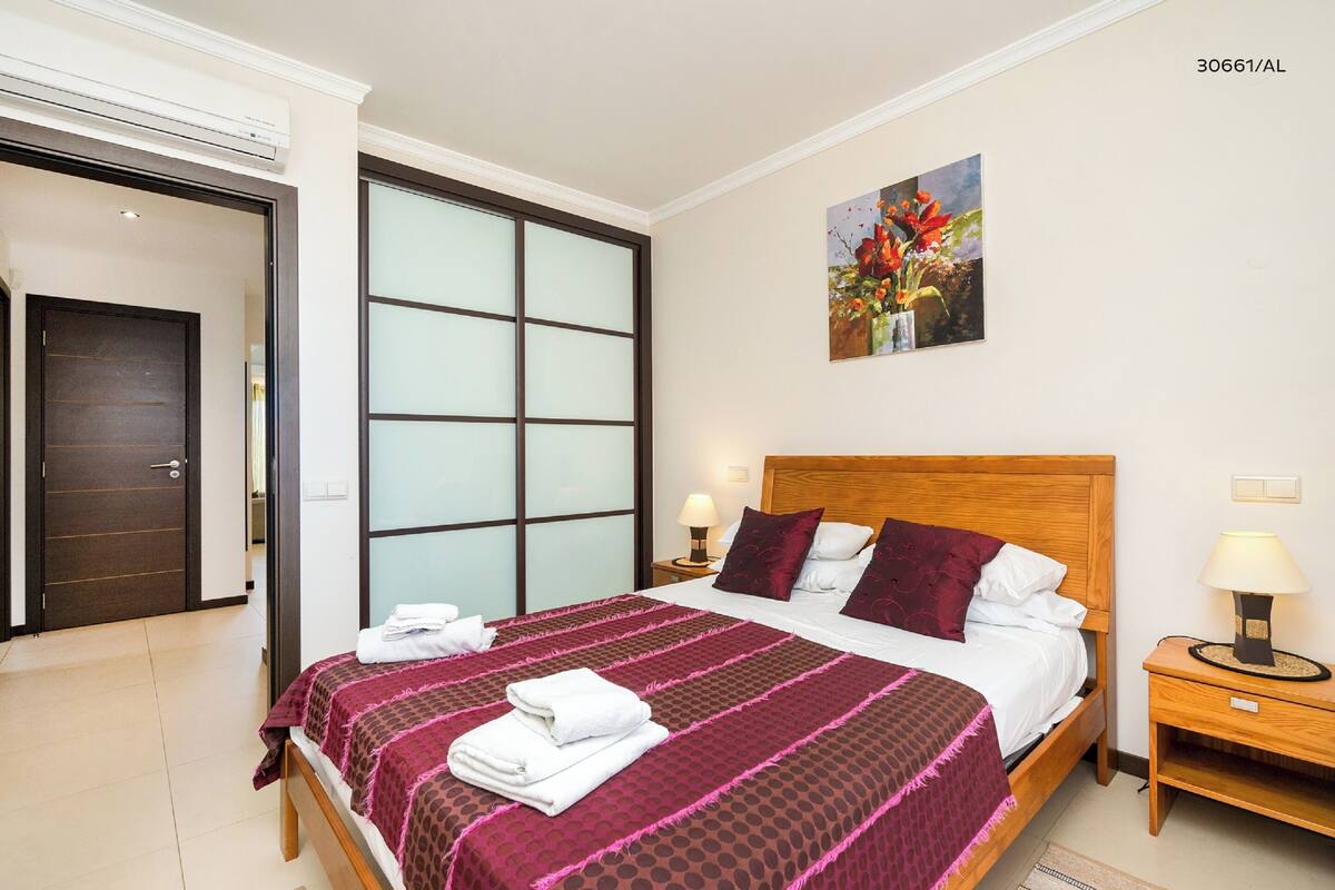 A hotel room with a large bed, a wooden headboard, a nightstand with a lamp, and a painting of flowers on the wall.