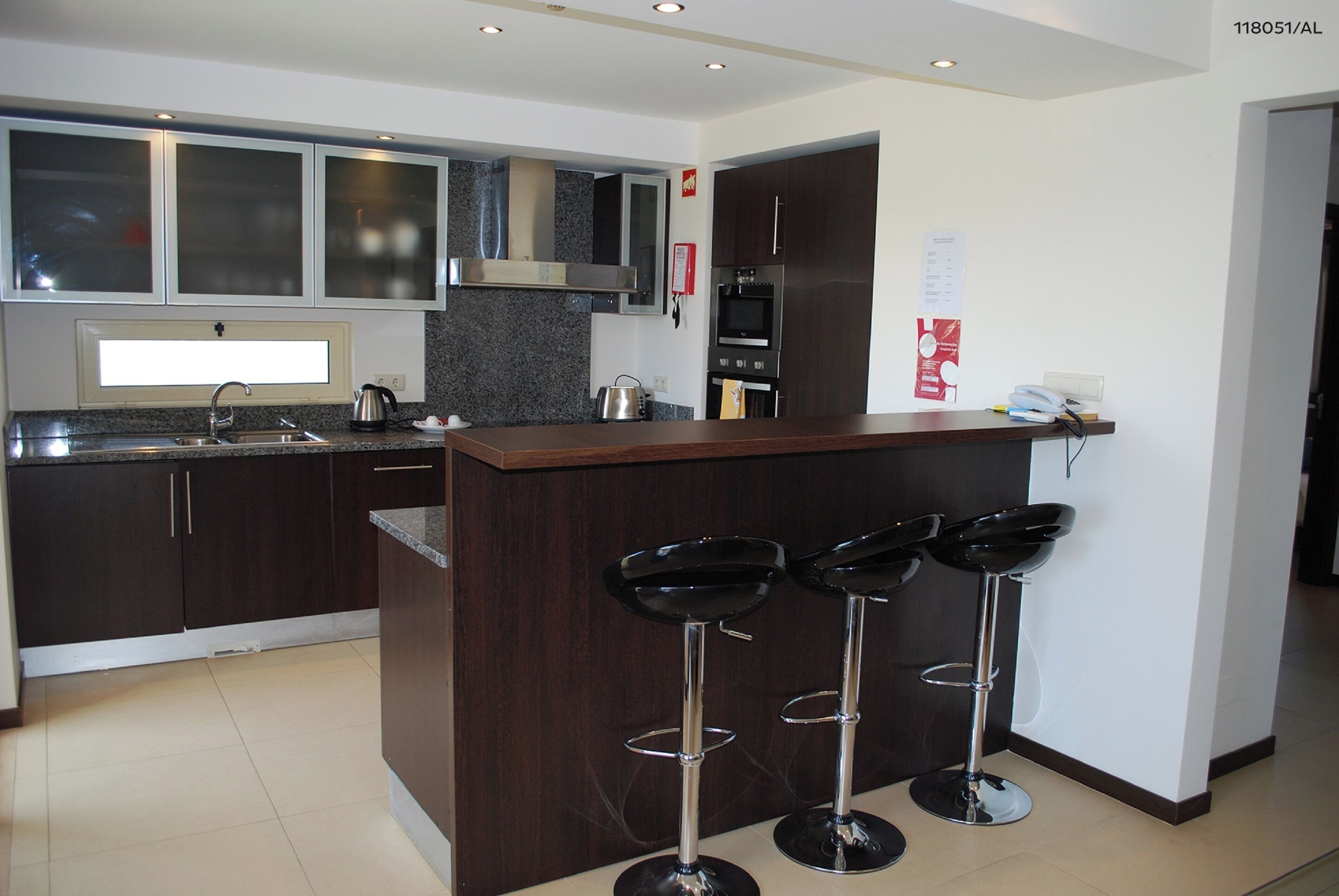 A modern kitchen with dark cabinetry, a granite countertop, a stainless steel sink, and a built-in microwave. There are three black bar stools with chrome legs.