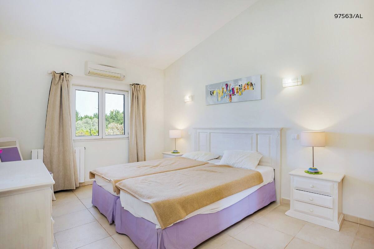 A bedroom with a double bed, a bedside table with a lamp, a window with curtains, and a painting on the wall.