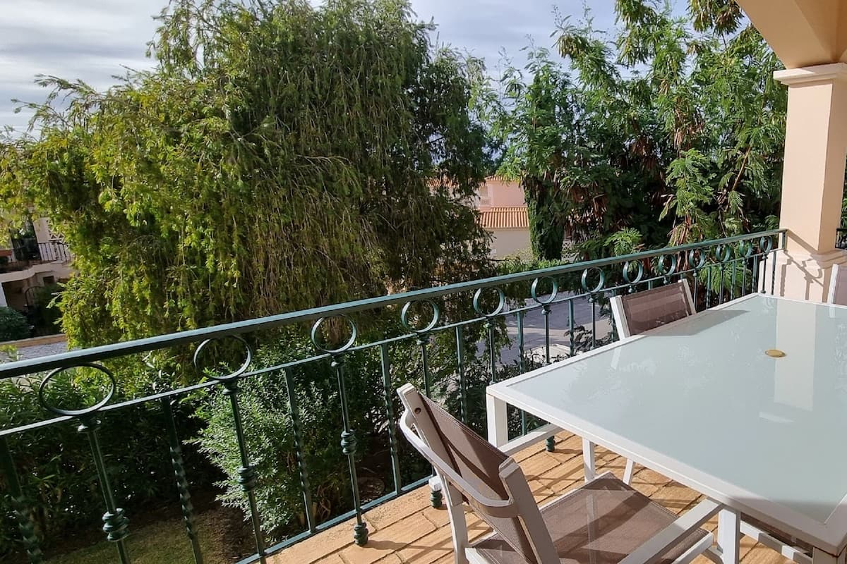 A balcony with a glass table and chairs, surrounded by greenery and a view of a building.