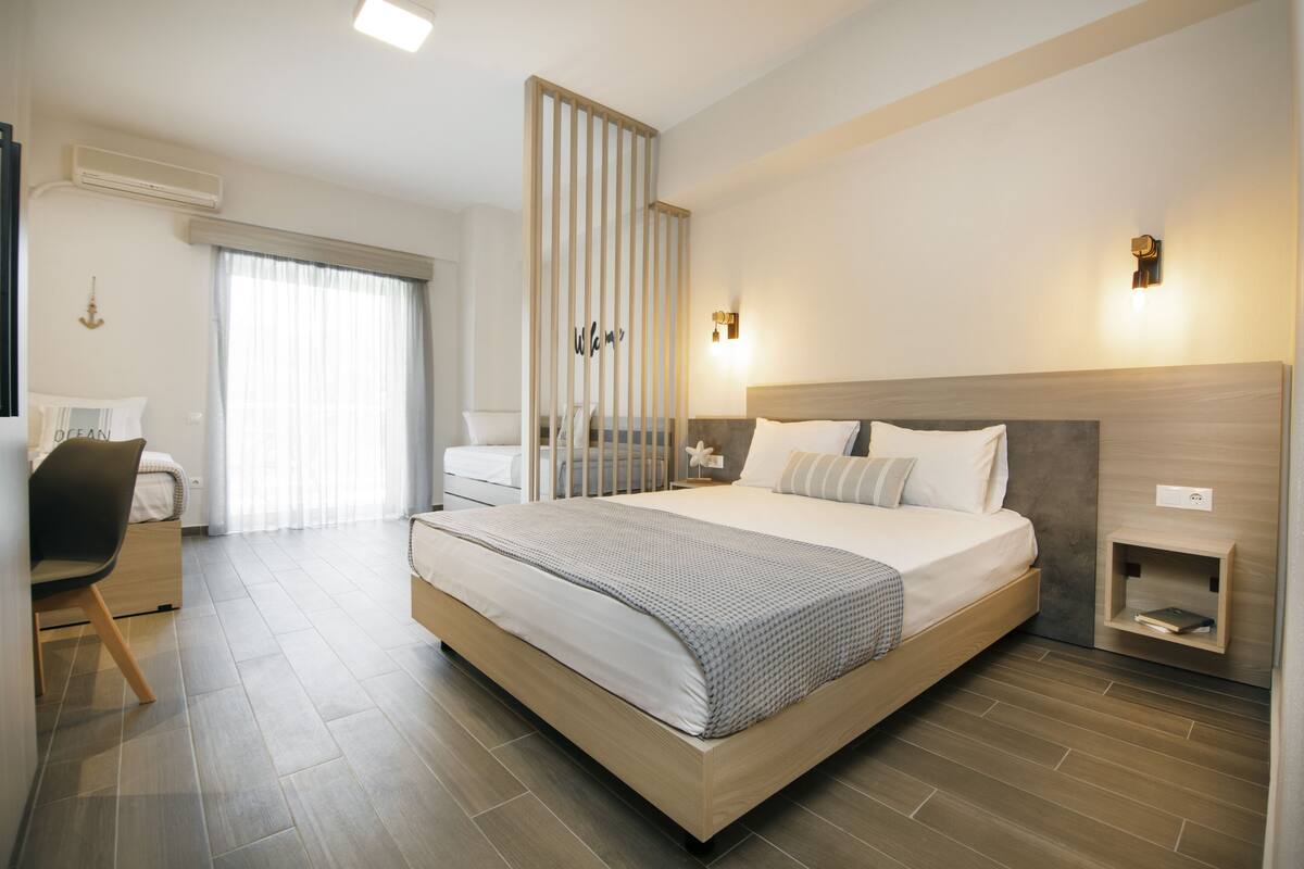 Habitación de hotel moderna con una cama grande, un escritorio y una silla. Cuenta con una ventana con cortinas, un aire acondicionado de pared y una pequeña mesita de noche con una lámpara.