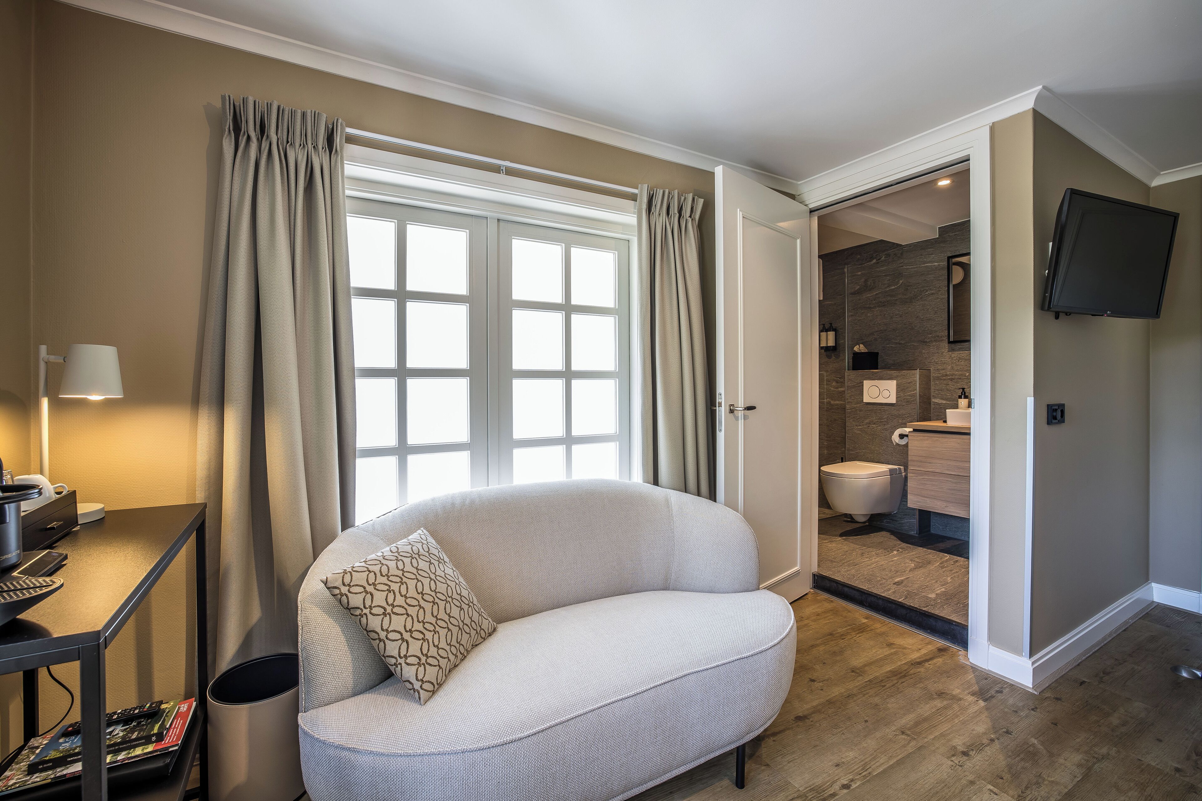 Moderni hotellihuone, jossa on suuri ikkuna, sohva, työpöytä lampun kanssa ja taulutelevisio.