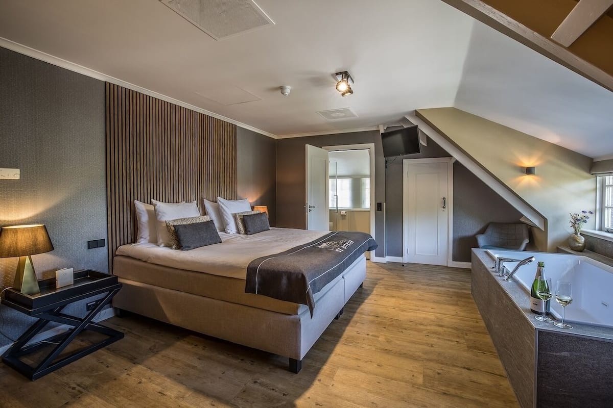 A modern bedroom with a large bed, a bedside table, a nightstand, a built-in bench, a bedside lamp, a wall-mounted shelf, and a window.