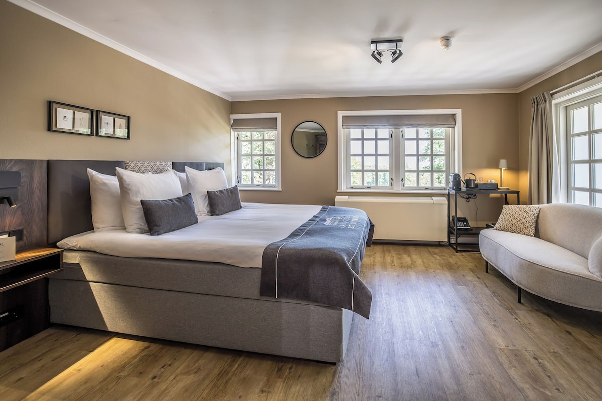 A bedroom with a large bed, a bedside table, a nightstand, a sofa, a window with curtains, and a wall-mounted mirror.