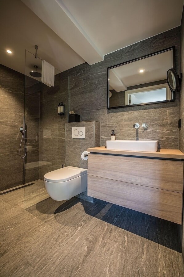 Un baño moderno con ducha envuelta en vidrio, inodoro blanco, tocador de madera con lavamanos y un espejo grande.