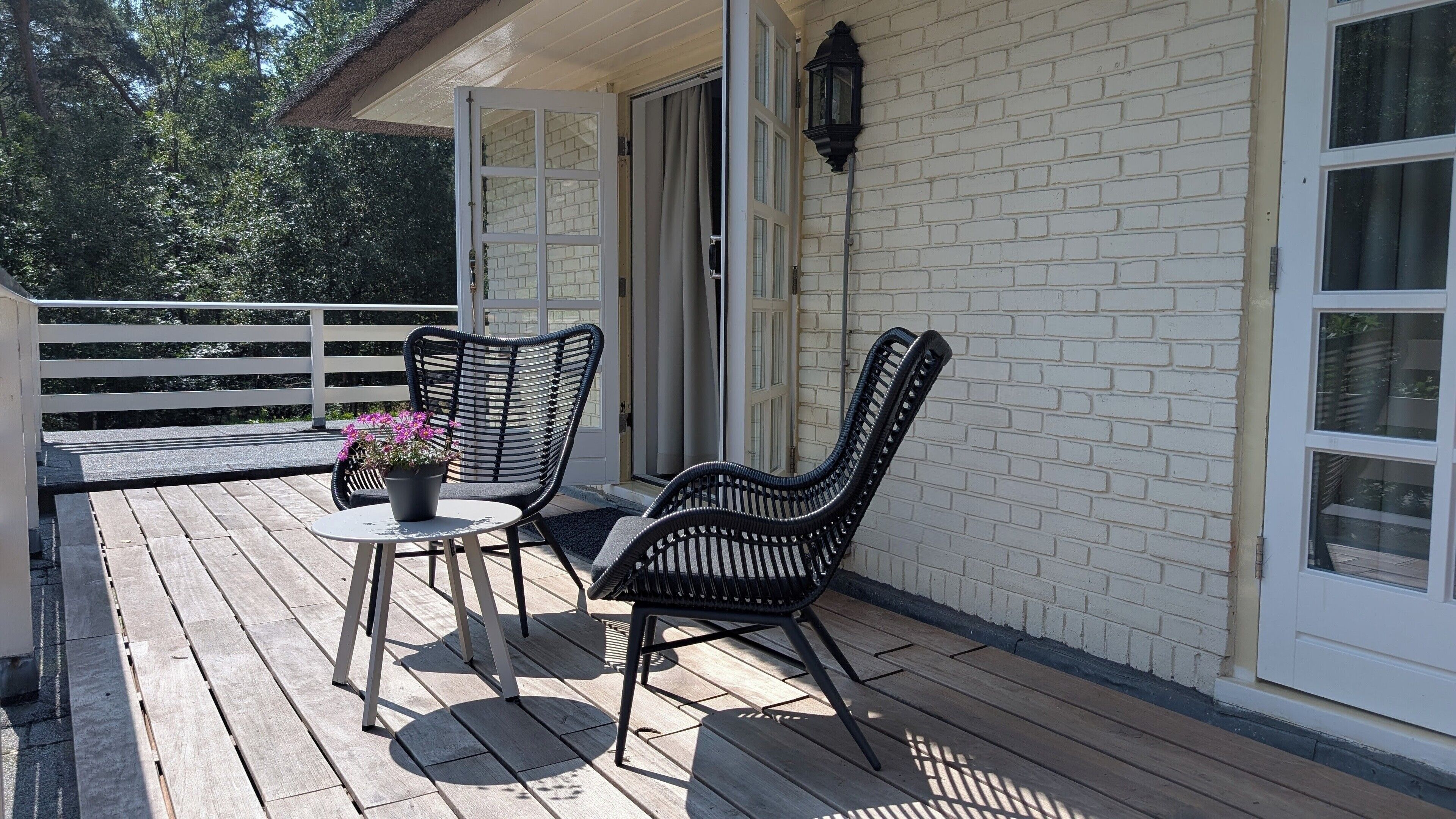 Een houten terras met twee zwarte rieten stoelen en een tafeltje met een plant in pot.