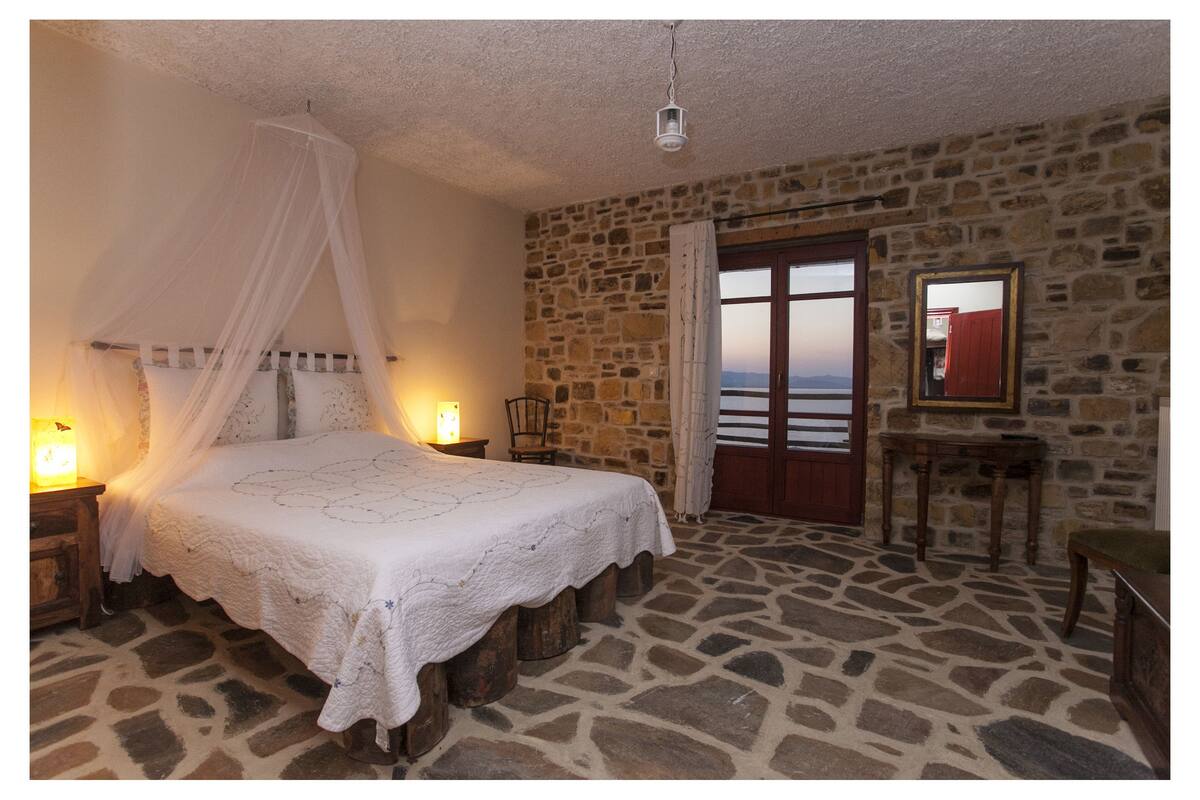 A bedroom with a stone floor, a bed with a canopy, a nightstand with a lamp, a wooden chair, a table with a mirror, and a view of the sea through a door.