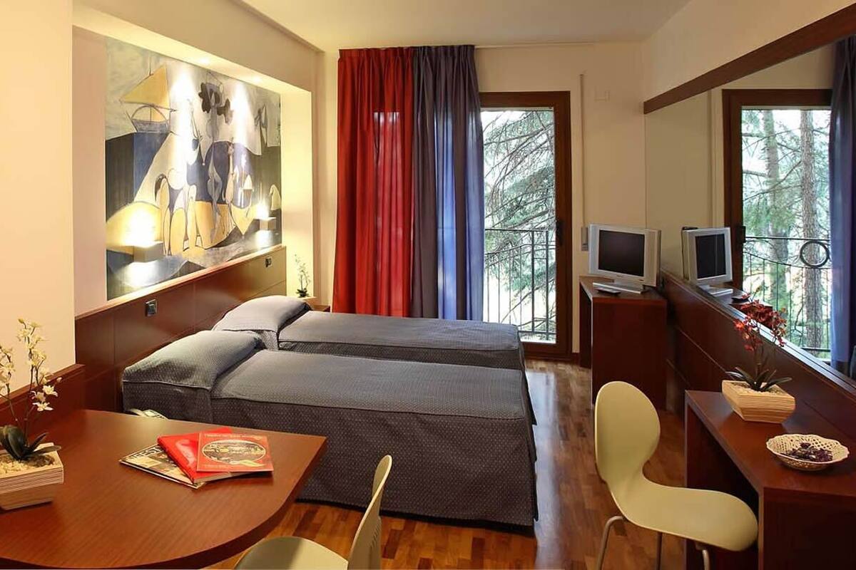 A compact hotel room with a bed, a desk with a chair, a television, and a window with curtains.