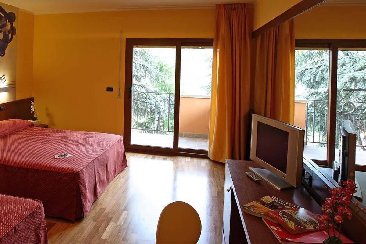 A hotel room with two beds, a TV, a balcony with curtains, and a large abstract painting on the wall.